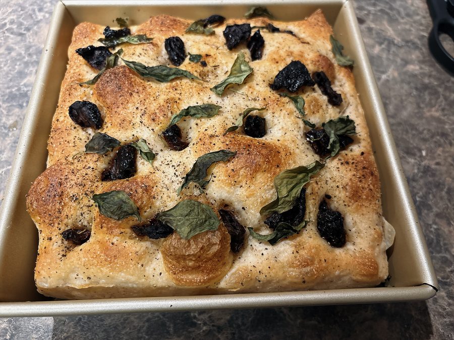 Focaccia baked in a square pan, dimpled and topped with herbs and dark olives
